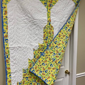May include: A white quilted baby blanket with a yellow and blue border. The blanket features a white quilted design of two wings. The border has a pattern of small animals and flowers.
