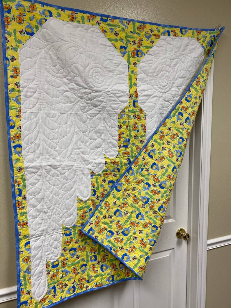 May include: A white quilted baby blanket with a yellow and blue border. The blanket features a white quilted design of two wings. The border has a pattern of small animals and flowers.