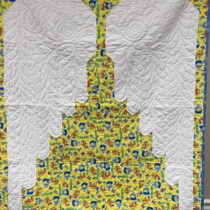 May include: A white quilted baby blanket with a yellow and blue fabric border. The blanket features a large white quilted design of angel wings.