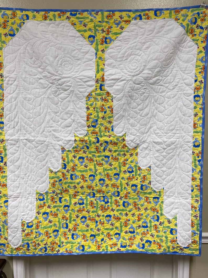 May include: A white quilted baby blanket with a yellow and blue fabric border. The blanket features a large white quilted design of angel wings.