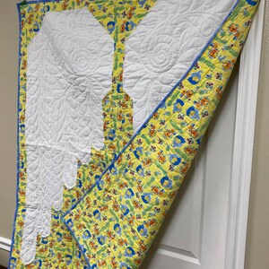 May include: A yellow and blue baby quilt with a white appliqu&eacute; of angel wings. The quilt is folded in half and hanging on a white door.