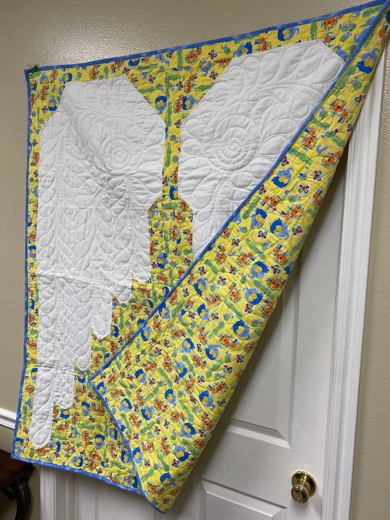 May include: A yellow and blue baby quilt with a white appliqu&eacute; of angel wings. The quilt is folded in half and hanging on a white door.