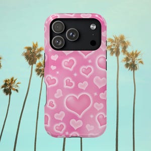 May include: Pink phone case with a pattern of hearts in varying sizes. The case has a glossy finish and a black camera module. The background features palm trees and a light blue sky.