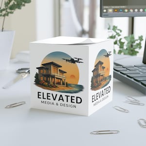 Puede incluir: Un bloc de notas blanco en forma de cubo con la inscripción "ELEVATED MEDIA & DESIGN". El diseño presenta una ilustración de una casa moderna, un dron y un atardecer. Varios clips plateados están dispersos alrededor del bloc.