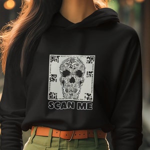 May include: A black hoodie featuring a white skull design made from a QR code. The text "SCAN ME" is printed beneath the skull. The model is wearing a brown belt and green trousers.