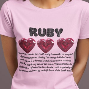 May include: Pink t-shirt with the word "RUBY" in black letters and three red ruby gemstones in the shape of hearts. The text below the gemstones reads "Its connection to the Earth, ruby is considered a crystal of grounding and vitality. Its energy is linked to the earth, since it is formed within rocks and is extracted from the depths of the earth's crust. This connection to the Earth is reflected in its red color, which symbolizes the primordial energy and life force of the Earth itself."