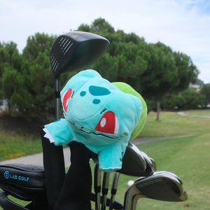 Op de afbeelding: Een zwarte golftas met een groene en blauwe pluche karakter-kopbedekking bevestigd aan een golfclub. De tas heeft de tekst "LAS GOLF" aan de zijkant.