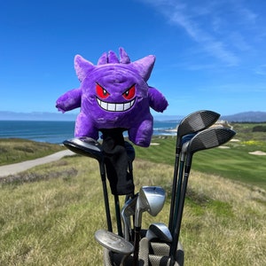 May include: A golf bag filled with golf clubs, topped with a plush purple Gengar headcover. The headcover has red eyes and a white grin. The bag is on a grassy hill overlooking the ocean and a golf course.
