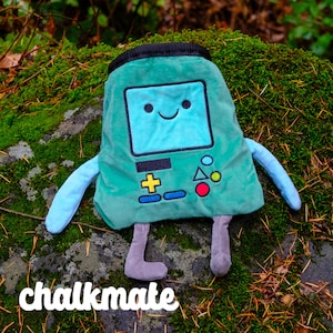 May include: A green plush toy shaped like a video game console with a blue screen and a smiling face. The toy has grey legs and arms and is sitting on a bed of moss.