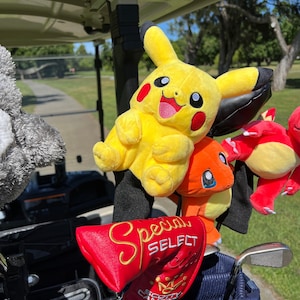 Può includere: Una sacca da golf con un copri-testa rosso che dice "Special Select" in lettere dorate. La sacca ha un design blu e nero ed è piena di mazze da golf. Ci sono diversi peluche attaccati alle mazze, tra cui un Pikachu giallo, un Eevee arancione e un Charizard rosso.