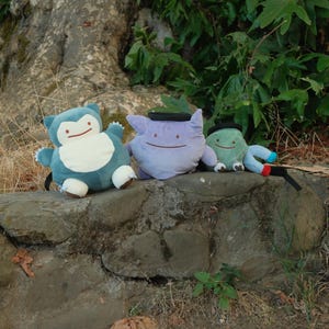 Pu&ograve; includere: Tre peluche, un Snorlax blu e bianco, un Gengar viola e bianco e un Bulbasaur verde e bianco, sono seduti su un muro di pietra.