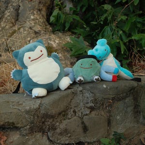 Pu&ograve; includere: Tre peluche, un Snorlax blu e bianco, un Bulbasaur verde e bianco e uno Squirtle blu e bianco, sono seduti su un muro di pietra.