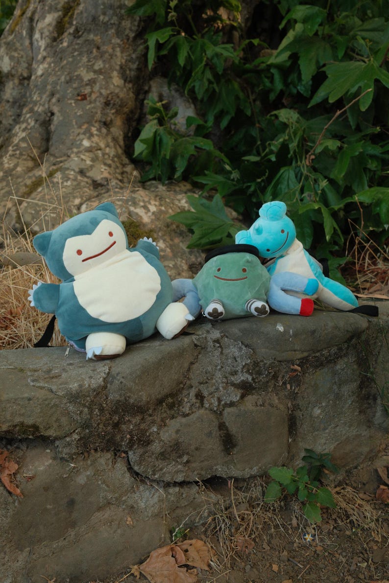 Pu&ograve; includere: Tre peluche, un Snorlax blu e bianco, un Bulbasaur verde e bianco e uno Squirtle blu e bianco, sono seduti su un muro di pietra.