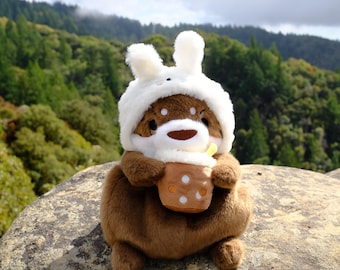 May include: A brown plush toy wearing a white bunny hat with floppy ears, holding a brown cup with white polka dots. The toy is sitting on a large, grey rock with green foliage in the background.