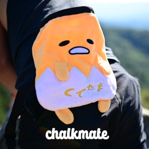 May include: A yellow and white plush character with a cute face and the Japanese text "Kute Tama" on the front. The character is wearing a black strap and is being held by a person.