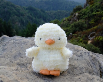 Pode incluir: Um brinquedo de pelúcia de pato branco e fofo com bico e pés laranja. O bicho de pelúcia está situado em um cenário de uma paisagem montanhosa e florestada. O brinquedo está sentado em uma grande rocha cinza.