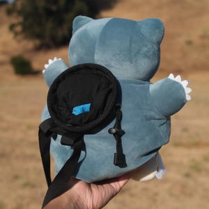 Pu&ograve; includere: Uno zaino di peluche blu a forma di personaggio Pok&eacute;mon Snorlax. Lo zaino ha una tasca nera sul retro con una zip blu.