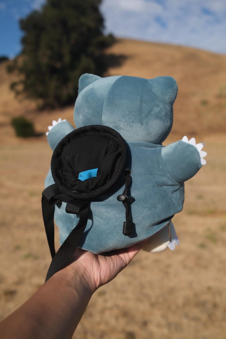 Pu&ograve; includere: Uno zaino di peluche blu a forma di personaggio Pok&eacute;mon Snorlax. Lo zaino ha una tasca nera sul retro con una zip blu.