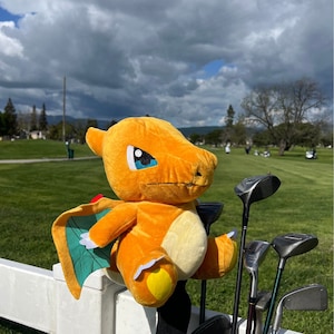 May include: A plush orange and green Charizard golf club headcover with a blue eye. The headcover is attached to a golf club in a golf bag.