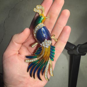 May include: A gold-toned peacock brooch with a vibrant design. The bird features a blue body, green and yellow tail feathers, and is adorned with clear crystals. The brooch is an elegant accessory.