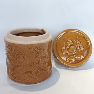 May include: A brown ceramic jar with a lid. The jar has a textured surface with a design of flames and clouds. The lid has a raised design of flames and clouds.