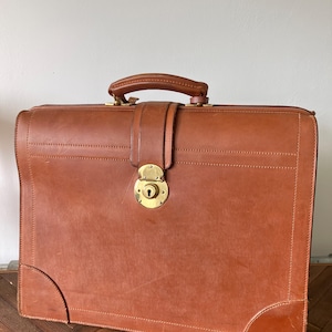 May include: A tan leather briefcase featuring a structured rectangular shape, a top handle, and a central vertical strap secured by a circular brass lock mechanism. The exterior displays visible stitching along the edges and reinforced corners.