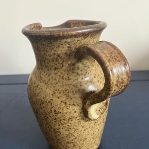 May include: A small, speckled ceramic jug with a curved handle. The jug is a warm tan colour with darker brown speckles and a brown rim. The handle is thick and rounded, and the jug has a slightly flared opening.