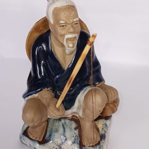 May include: A ceramic figurine of a man with a white beard and a blue robe, sitting on a rock and holding a fishing rod. He is wearing a straw hat and has a relaxed expression on his face.