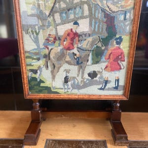 May include: A framed needlepoint scene showing figures in red coats on horseback with dogs, set against a building and trees. The frame and stand are made of dark wood, creating a vintage aesthetic.