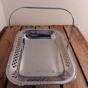 May include: A polished, silver serving tray with a handle. The rectangular tray features decorative cut-out patterns around the edges and a curved handle. It is displayed on a wooden surface.