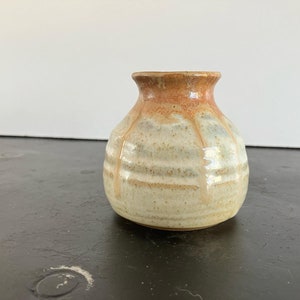 May include: A small, round, ceramic vase with a light brown glaze and a darker brown rim. The vase has a subtle, textured surface.