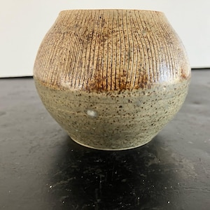 May include: A small, round, ceramic vase with a textured, brown and green glaze. The vase has a wide opening and a rounded bottom.
