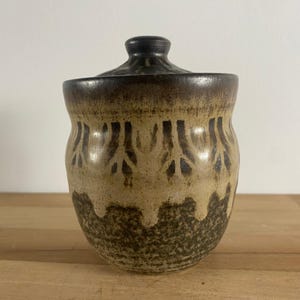 May include: A brown and tan ceramic jar with a lid. The jar has a decorative pattern of brown lines and shapes.