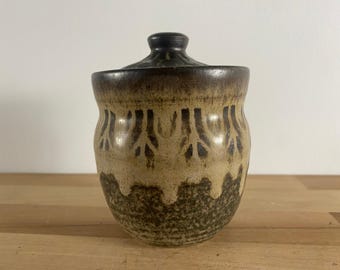 May include: A brown and tan ceramic jar with a lid. The jar has a decorative pattern of brown lines and shapes.