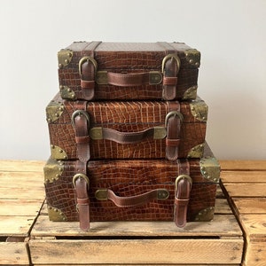 May include: Three brown faux-leather suitcases stacked on top of each other, featuring brass corner protectors and buckles. The cases have a crocodile skin texture and leather handles. They are placed on a wooden crate.