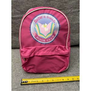 Puede incluir: Una mochila rosa con ribete blanco y un bolsillo delantero. La mochila presenta un emblema circular con el texto "Barbie & friends BOOK CLUB" y un gráfico de un libro abierto con corazones.