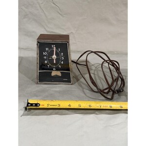 May include: A vintage black and brown electric clock with a white face and a brown power cord. The clock face has white numbers and hands. A yellow measuring tape is in the foreground.