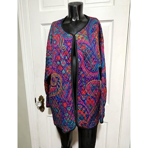 May include: A quilted open-front jacket featuring a vibrant paisley pattern in shades of purple, blue, pink, and red. The garment has long sleeves, a rounded neckline, and black trim along the edges, providing a structured silhouette.