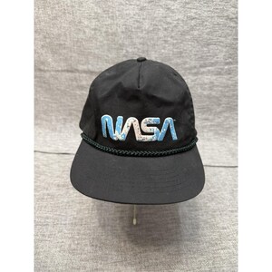 May include: Black baseball cap featuring the NASA logo in white and blue. The cap has a curved peak and a dark green rope detail around the base. The NASA logo is in a stylized font.