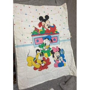 Könnte beinhalten: Weiße Babydecke mit Abbildungen von Micky Maus, Minnie Maus, Donald Duck und Pluto. Die Decke hat einen blauen Streifen und bunte Tupfen. Die Zeichentrickfiguren sind in leuchtenden Farben gehalten, mit einem Farbschema aus Rot, Blau, Gelb und Grün.
