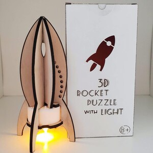 Peut inclure: Puzzle de fusée 3D en bois avec lumière. La fusée est en bois brun clair avec des accents noirs et une lumière jaune vif à la base. La boîte à côté porte un graphique de fusée rouge et le texte "3D ROCKET PUZZLE WITH LIGHT".