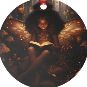May include: Round Christmas ornament featuring a fairy with butterfly wings reading a book in a forest setting. The fairy has dark curly hair and is wearing a crown. The ornament has a red ribbon for hanging.