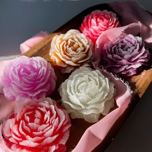 May include: Six handmade soap flowers in a variety of colors, including pink, purple, white, and orange, arranged on a wooden tray with a pink fabric background.