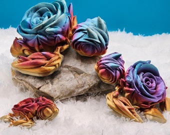 May include: Five iridescent rose-shaped sculptures in shades of blue, purple, and gold. The sculptures are arranged on a rock and a white fluffy surface.