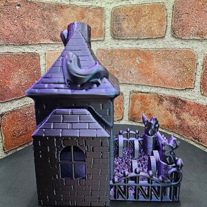 May include: A 3D printed purple and black haunted house with a black cat on the roof and a black fence with three black cats in the yard.