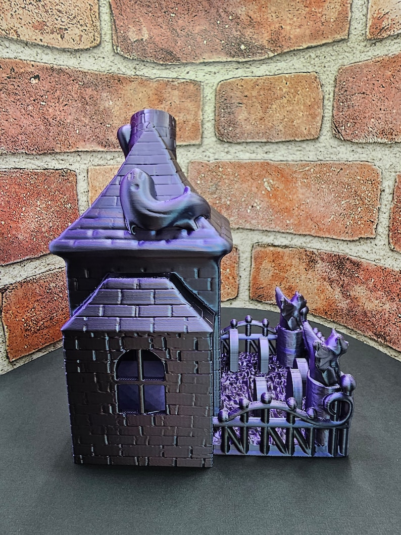 May include: A 3D printed purple and black haunted house with a black cat on the roof and a black fence with three black cats in the yard.