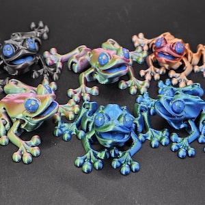 May include: Five small frog figurines in different colors, including black, green, blue, and iridescent. The frogs are all in a smiling pose with their mouths open.