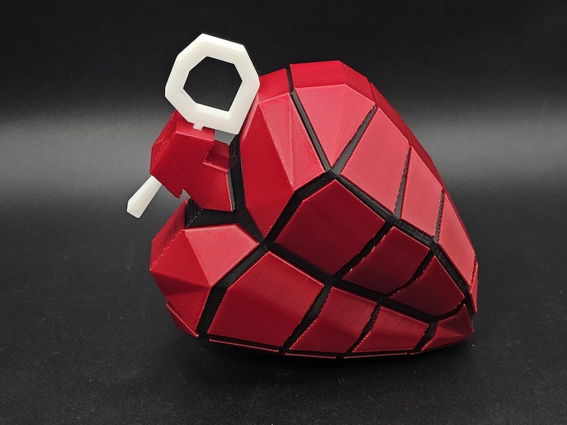 May include: A red and black heart-shaped grenade with a white pull ring. The grenade is made of a 3D printed material and has a geometric design.