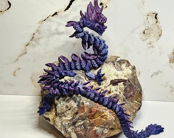 May include: A purple and blue articulated dragon sculpture with a detailed design. The dragon is perched on a natural stone with a textured surface. The sculpture is approximately 25 cm long.