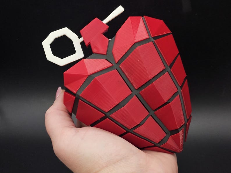 May include: A red and black heart-shaped grenade with a white pin. The grenade is made of a 3D printed material and has a textured surface.